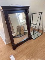 Wooden framed mirror with carved edges standing on floor next to metal framed mirror shelf with reflective base