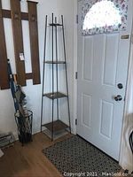 Full view of the tall metal plant stand near a door, showing its slim profile and four tile shelves.