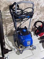 Front angle shot showing the full pressure washer unit with hose wrapped around the handle, plastic lance attached, two wheels at base, and control dial on top.