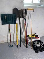 Photo of various shovels, snow shovel, paint roller with extension pole, and a crate of tools including plumbing equipment, measuring tape, saws, and trimmers against a white basement wall.