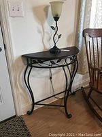 Side view of the half-moon shaped wood top side table with black metal base and leaf design near the front. Table stands on four curved legs.