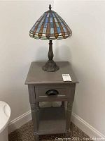 Full view of Tiffany style table lamp placed on wooden nightstand with drawer and lower shelf, lamp has colorful stained glass shade and ornate bronze base