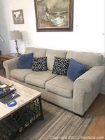 Front and side view of beige three cushion sofa with four throw pillows on a striped area rug, wooden side table and lamp visible.