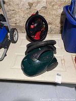 Two vacuum cleaners on a table near basement wall: red Hyper Tough shop vac and green Kenmore canister vac.