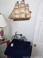 Wall art set with sailing ship mounted above two metal bicycle wall art pieces on a white wall, with a lamp and end table nearby.