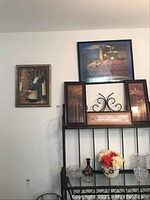 Wide shot showing 5 pieces of wall art on a white wall above a metal stand with glass items and flowers.