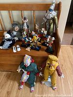 Photo shows complete assortment of clown figurines and dolls arranged on wooden bench, includes porcelain and fabric items of various sizes.