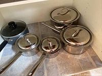 Photo showing five assorted pots placed on a floor with tile pattern. Four pots are stainless steel with shiny lids, one pot is dark grey with non-stick coating and a glass lid with black knob.