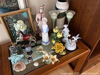 Photo showing miscellaneous decor items on mirrored surface atop wooden dresser, including porcelain figurines, glass calla lily vases, ceramic flowers, and small decorative pieces.