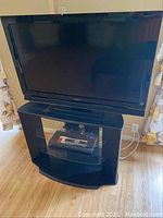 Front view of Sony 40-inch TV mounted on black glass shelf stand in room setting, showing TV screen and stand with glass shelves.