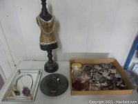 Jewelry lot showing black mannequin dress form jewelry display, small round pedestal display, mirrored tray with assorted jewelry, and cardboard box filled with various jewelry pieces including necklaces, bracelets, pins, and earrings.