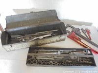 Full view of vintage light grey metal toolbox with removable tray inside containing wrenches and socket wrenches along with larger pipe wrenches outside the toolbox.