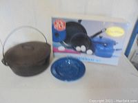 Photo showing Lodge cast iron Dutch oven with lid and handle beside the Omni 7-piece cookware set box and one enamelware plate.