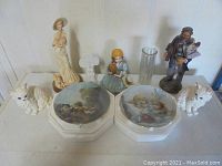 Wide shot showing the main ceramic figurines and two dog collector plates with a glass vase against a white wall.