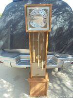 Full view of the Bentley IX grandfather clock showing the wooden body, mirrored front with pendulum and weights hanging, against a vehicle background.