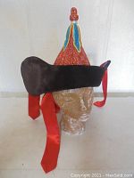 Side and front angled view of the Mongolian hat showing the conical shape, colorful patterned fabric, black fur brim, and red satin ribbons.