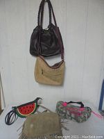 Five purses displayed, two hanging on the wall (dark brown leather and tan suede), three on a surface (watermelon-themed small purse, fringe tan bag, camouflage pink LOVE bag)