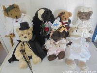 Seven teddy bears sitting on a white cloth background, dressed in various costumes including wedding attire and nurse-like uniform.