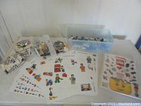 Photo showing four small unopened Lego bags numbered 3 and 4 with various bricks, a plastic tub with loose pieces, seven Lego minifigures, and printed Lego sticker sheets.