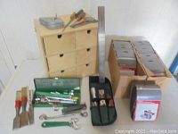 Photo showing wooden storage box with six drawers, metal ruler, socket set case, chisels, adjustable wrench, and two boxes of sanding belts.