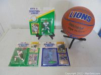 Photo showing three Starting Lineup sports figures new in packaging with Bo Jackson, Barry Sanders, and Deion Sanders, alongside signed Detroit Lions basketball on display stand.
