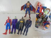 Group of superhero action figures including Spider-Man, Joker, Batman, Nightwing, Superman, Wonder Woman and others in a white bin and on a table