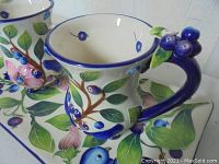 Close-up photo of one ceramic cup showing blue berry decorated handle, painted leaves and blue rim