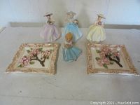 Full lot of four ceramic lady figurines and two floral relief plates, shown on a table.