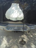 Full view of the vintage floor lamp showing the white fabric lampshade with fringe and the ornate metal body against a vehicle background.