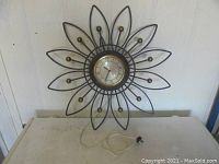 Full view of the vintage metal wall clock showing the black metal sunburst frame with brass balls and attached electric cord on a white surface.