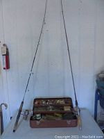 Two fishing poles with cork handles and attached reels standing against a wall behind a reddish metal tackle box laid open with various tackle inside.