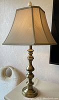 Full view of the brass lamp with beige fabric lampshade set on table next to cream upholstered chair.