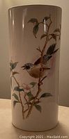 Side view of a tall cylindrical porcelain vase showing a hand painted bird perched on a flowering branch with green leaves and pink flowers. Glossy finish with detailed naturalistic painting.