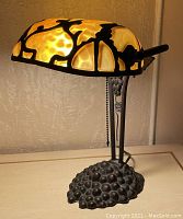 Desk lamp lit up, showing yellow glass shade and painted bronze grape vine design base and support.