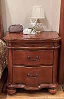 Front view of one dark wood bedside table showing two drawers with metal handles, curved top edge, and rounded bun feet. Decorative molding along drawer fronts and bottom skirt.