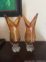 Pair of orange Murano glass vases with clear glass pedestal bases on a dark wooden surface under painting, showing flared organic shapes and translucent orange color.