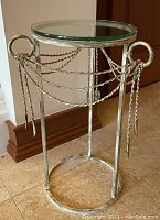 Side table with round glass top, metal base with twisted rope and leaf accents, distressed silver finish.