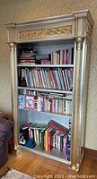 Front view of the tall Italian bookshelf showing three shelves filled with books and ornate gold trim and columns with a silver base finish.