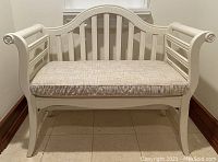 Front view of white wooden bench with upholstered seat cushion, showing curved slatted back and rolled arms.
