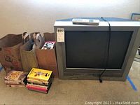 Silver Toshiba MW24FM3 CRT TV with built-in VHS player and remote on top; VHS movie tapes in their cases stacked beside the TV; brown paper bags behind the tapes for carrying or storage.