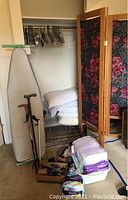 Wide photo showing piled linens and pillows on wire shelf, ironing board leaning against closet wall, canes in holder, box with dumbbells and shoe polish items on floor, and floral fabric room divider panels.