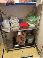 Metal shelving with stacked plastic food storage containers, cookpot placed underneath and the propane stove on the floor below shelf