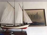 View of the hand built sloop model with sails and stand in front of the framed art. The sparkplug airplane is visible under the boat stand.