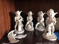 Four white Dresden porcelain cherub figurines displayed on a wooden shelf. Each cherub is in a different pose with detailed wings and decorative floral bases. One separate wing piece is also present.
