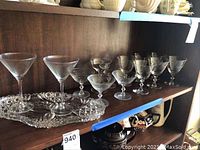 Photo of two taller etched cordial glasses and six smaller etched cordial glasses on an etched serving tray, on a wooden shelf.