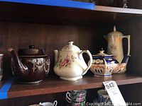 Shelf with four ceramic teapots showing varying colors and floral/ornate decorations from dark brown to cream and blue.