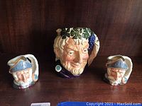 Front view of the three Royal Doulton character jug and cups, showing the large character jug with facial details and grapevine decoration and two smaller character cups with blue hats.