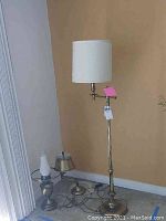 Photo of three vintage styled brass lamps against a beige wall: one floor lamp with a white fabric shade and two desk lamps with matching white shades beside it, showing cords and tarnished brass bases.