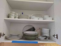 View of assorted dishware on two shelves inside a cabinet including floral tea cups, stacks of plates with green rims and white bowls, clear glass plates and bowls.