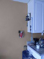 Wall corner with hanging medium metal cow bell and wreath below it, kettle and coffee items on countertop to right.